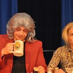 Zwei Frauen sitzen an einem Tisch, eine mit grauen Haaren, die eine Kaffeetasse hält, und die andere mit blonden Haaren in einem Leopardenmusterhemd. Die Frau mit grauen Haaren lächelt.