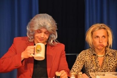 Zwei Frauen sitzen an einem Tisch, eine mit grauen Haaren, die eine Kaffeetasse hält, und die andere mit blonden Haaren in einem Leopardenmusterhemd. Die Frau mit grauen Haaren lächelt.