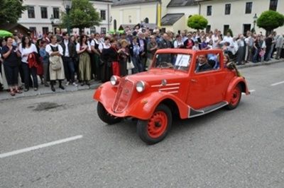 Ein altes rotes Auto fährt mit einer Menschenmenge, die zusieht, die Straße hinunter. Das Auto hat ein klassisches Design mit roten Rädern und einem Dach. Die Menschen sind in verschiedene Kleidung gekleidet, einige halten Regenschirme.