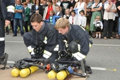 Zwei Feuerwehrleute in voller Ausrüstung knien auf der Straße und demonstrieren Ausrüstung, während die Leute hinter ihnen stehen und zusehen. Die Feuerwehrleute sind neben gelben Zylindern.