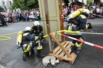 Drei Feuerwehrleute in voller Ausrüstung arbeiten an einer Tür, während eine Menge zuschaut. Einer hält einen Vorschlaghammer, ein anderer eine Seile und der dritte steht auf einer Holzplattform. Die Szene spielt sich auf einer Straße mit Zuschauern und Rettungsfahrzeugen ab.