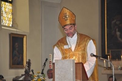 Ein Priester in goldener Stola und Mitra steht an einem Rednerpult mit Mikrofon und hält eine Rede. Dahinter befinden sich Kerzen auf einem Regal, ein Gemälde an der Wand und eine Statue.