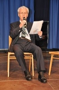 Ein älterer Mann in Anzug und Krawatte sitzt auf einem Holzstuhl auf der Bühne, hält ein Mikrofon und ein Stück Papier in der Hand und hält wahrscheinlich eine Rede.