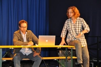 Ein Mann und eine Frau stehen auf der Bühne. Der Mann sitzt auf einer Bank, hält einen Stift und schreibt. Die Frau steht neben einem Tisch mit einem Laptop. Ein blauer Vorhang im Hintergrund.