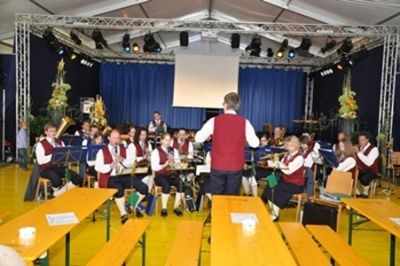 Eine Band spielt auf einer Bühne in einem Zelt. Die Musiker sitzen mit ihren Instrumenten auf Bänken. Ein Mann steht vor ihnen und leitet die Band. Scheinwerfer sind an der Decke montiert und eine Leinwand befindet sich im Hintergrund.