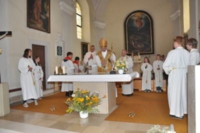 Eine Gruppe von Menschen in weißen Gewändern steht um einen Altar mit Blumenarrangement und Kerzen, vor ihnen ein Priester.