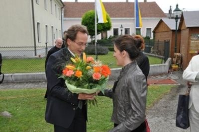 Ein Mann in einem Anzug überreicht einer Frau in einer grauen Jacke einen Blumenstrauß. Sie stehen auf einem Rasen. Hinter ihnen befinden sich Gebäude und ein Zaun.