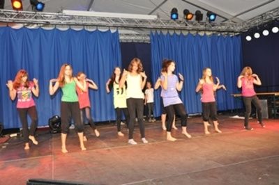Eine Gruppe von Frauen in verschiedenen farbigen Oberteilen und Leggings führt eine Tanzroutine auf einer Bühne mit blauen Vorhängen und Deckenbeleuchtung auf.