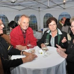 Eine Gruppe von Menschen, darunter drei Frauen und zwei Männer, versammelt sich unter einem Zelt um einen Tisch und hält Weingläser in der Hand.