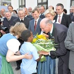 Ein älterer Mann erhält Blumen von einem Jungen in traditioneller Kleidung. Hinter ihnen beobachten mehrere Menschen, darunter ein Mann in Anzug und Krawatte.
