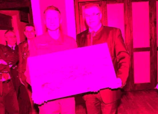 Ein Mann in Uniform hält ein großes rosa Plakat, während ein anderer Mann in Anzug neben ihm steht, beide sehen in die Kamera. Dahinter stehen weitere Männer in Uniformen in einem rosa beleuchteten Raum.