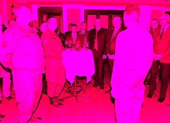 Eine formelle Zusammenkunft mit Menschen in Anzügen, einige in Militäruniformen, stehen um einen Tisch mit Getränken und Gläsern herum. Der Raum ist in einem pinken Licht erleuchtet.