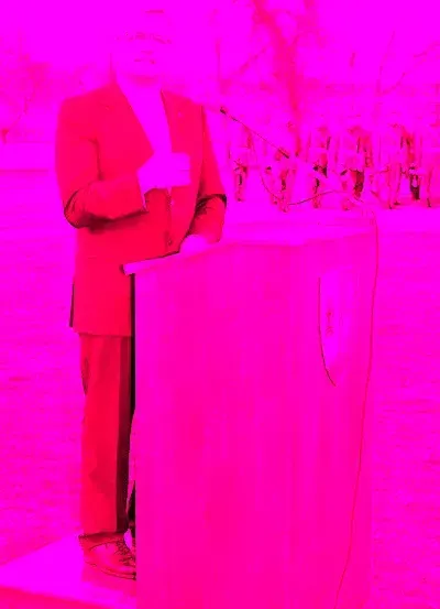 Eine Person steht an einem Rednerpult mit Mikrofon, gekleidet in einem roten Anzug, auf einer rosa getönten Bühne mit verschwommenen Figuren im Hintergrund.