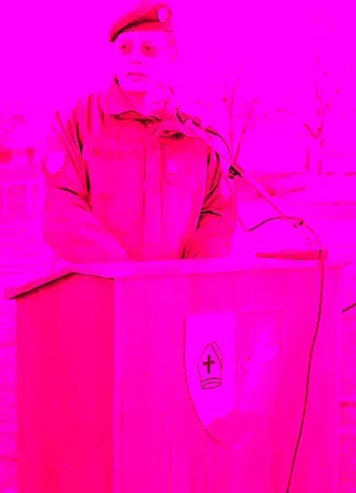 Ein Mann in einem roten Uniform und Hut steht an einem Rednerpult mit einem Mikrofon. Das Rednerpult ist rot und hat ein Kreuzsymbol darauf. Der Hintergrund ist pink.