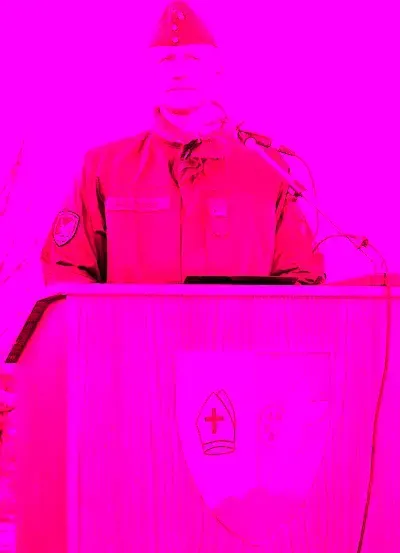 Ein Mann in Uniform steht an einem Podium mit Mikrofon, davor ein rotes Kreuz.