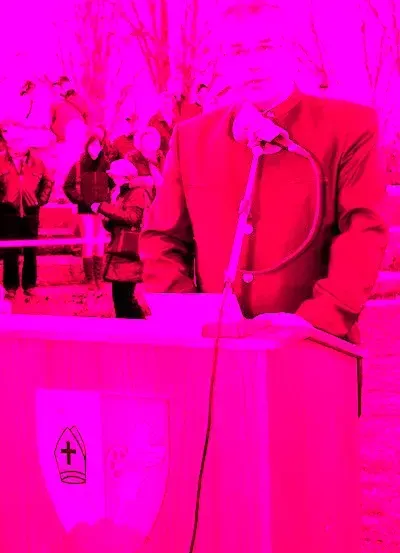 Ein Mann in einem roten klerikale Gewand steht an einem Rednerpult mit einem Mikrofon, möglicherweise hält er eine Rede. Dahinter steht eine Menschenmenge, die zusieht.