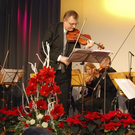 Ein Geiger führt ein kleines Orchester auf der Bühne an, mit anderen Musikern dahinter. Die Bühne ist mit roten Blumen geschmückt.