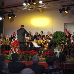 Ein klassisches Orchester spielt auf einer Bühne mit roten Christsternen und einem Dirigenten. Das Publikum sitzt vor der Bühne.