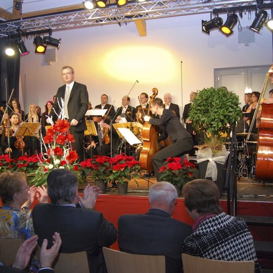 Ein klassisches Orchester tritt auf einer Bühne mit roten Blumen auf. Ein Publikum sieht ihnen zu.