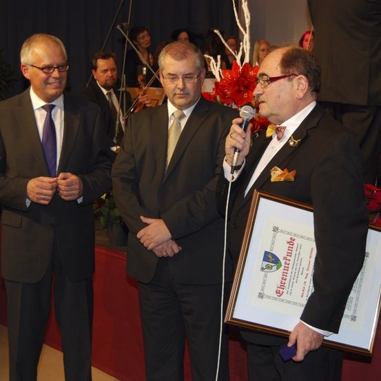 Drei Männer in Anzügen stehen auf einer Bühne, einer hält ein gerahmtes Zertifikat und ein Mikrofon. Ein roter Blumenstrauß ist im Hintergrund.
