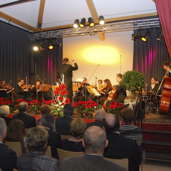 Ein klassisches Orchester spielt auf einer Bühne mit roten Blumen, unter Bühnenbeleuchtung, vor einem sitzenden Publikum.