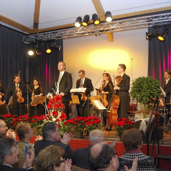 Ein klassisches Musikensemble spielt auf der Bühne mit roten Blumen, unter Bühnenlichtern, vor einem Publikum.