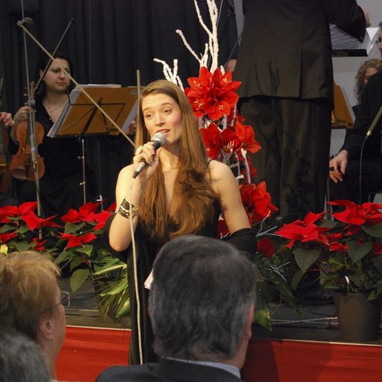 Eine Frau singt auf der Bühne und hält ein Mikrofon. Hinter ihr befinden sich ein Geiger und ein Cellist. Vor ihr sitzt ein Publikum. Die Bühne ist mit roten Blumen geschmückt.