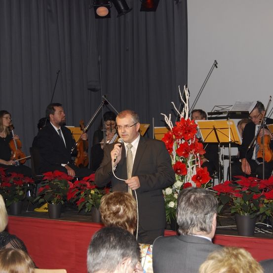 Ein Mann steht auf der Bühne und spricht in ein Mikrofon vor einem sitzenden Publikum. Hinter ihm spielt ein kleines Orchester, und die Bühne ist mit roten Blumen geschmückt.