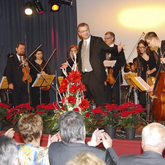 Ein Mann in Anzug dirigiert ein Streichorchester auf der Bühne mit roten Blumen. Das Publikum klatscht.