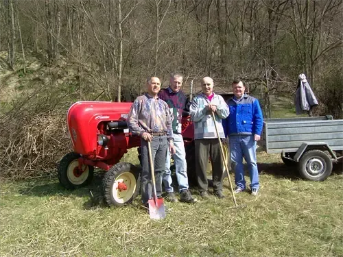 Vier Männer stehen neben einem roten Traktor mit Anhänger. Sie halten alle Schaufeln. Der Hintergrund zeigt kahle Bäume.