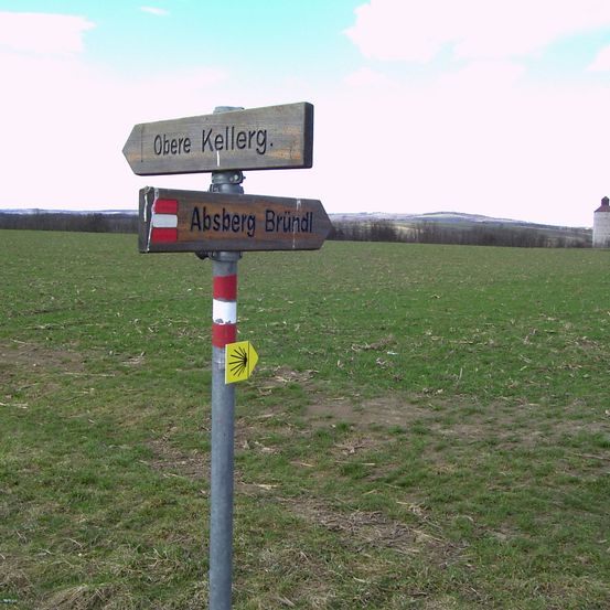 Ein Wegweiser mit zwei Holzschildern steht auf einer Wiese. Das oberste Schild zeigt 'Obere Kellerg'. Das untere Schild zeigt 'Absberg Brundl'. In der Ferne steht ein Turm.