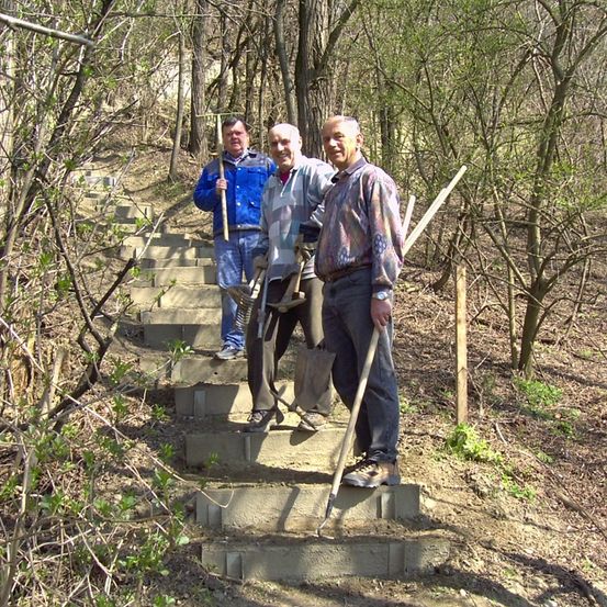 Drei Männer stehen auf einer Reihe von Betonstufen im Freien. Sie halten Schaufeln in den Händen und sind wahrscheinlich mit Bau- oder Wartungsarbeiten beschäftigt.