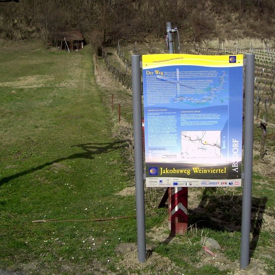 Ein Wegweiser für den Jakobsweg Weinviertel befindet sich auf einer grasbewachsenen Fläche in der Nähe eines Weinbergs. Der Schild hat eine Karte und etwas Text und ist an Metallpfosten befestigt.