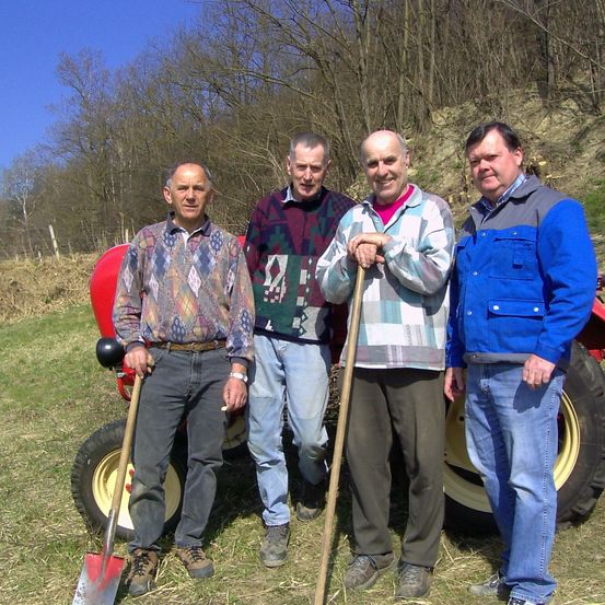 Vier Männer stehen in einem Feld mit einem Traktor, jeder hält eine Schaufel und einen Stock. Sie tragen Freizeitkleidung, einer trägt einen Gürtel. Der Traktor ist rot mit gelben Rädern. Hinter ihnen stehen Bäume und Büsche.