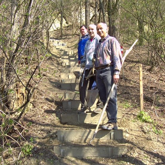 Drei Männer stehen auf einer Steintreppe im Wald und halten Schaufeln. Ein Mann lächelt in die Kamera, während die anderen beiden wegschauen.