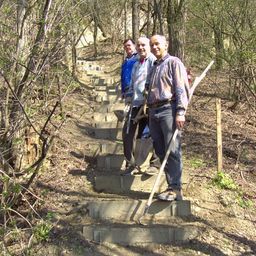 Drei Männer stehen auf einer Steintreppe im Wald und halten Schaufeln. Ein Mann lächelt in die Kamera, während die anderen beiden wegschauen.