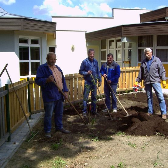 Vier Männer arbeiten in einem Garten und tragen Handschuhe. Ein Mann hält eine Harke und andere halten Schaufeln. Sie stehen in einem Erdbereich mit einem Holzzaun hinter ihnen.