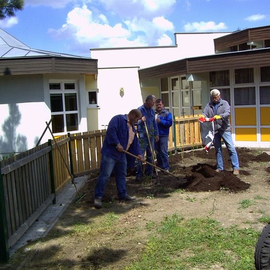 Vier Männer arbeiten in einem Garten vor einem Gebäude. Sie benutzen Rechen und Schaufeln zum Graben und Pflanzen. Der Bereich ist von einem Holzzaun umgeben.