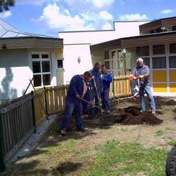 Vier Männer arbeiten in einem Garten vor einem Gebäude. Sie benutzen Rechen und Schaufeln zum Graben und Pflanzen. Der Bereich ist von einem Holzzaun umgeben.