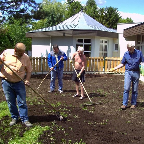Vier Männer arbeiten in einem Garten und benutzen Rechen. Einer trägt Handschuhe, ein anderer eine Jacke. Ein Gebäude mit einem Kuppeldach und einem Zaun befindet sich hinter ihnen.
