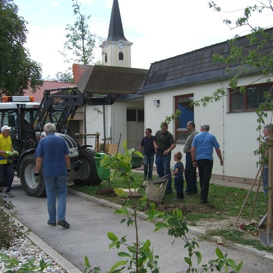 Eine Gruppe von Menschen versammelt sich um ein Gebäude mit einem Turm, und sie sind mit einer Aktivität beschäftigt. Ein Baufahrzeug ist in der Nähe geparkt. Sie tragen alle Freizeitkleidung. Ein Kind hält eine Schaufel.