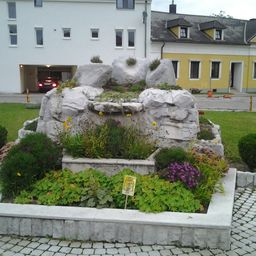 Ein Brunnen, umgeben von Felsen und verschiedenen Pflanzen, einschließlich gelber und lila Blumen, befindet sich vor einem Gebäude mit einer Garage.