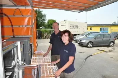 Zwei Personen stehen neben einem Food-Truck. Sie lächeln und scheinen für ein Foto zu posieren. Der Food-Truck hat ein Verdeck und eine Aufbewahrungsbox. Im Hintergrund sind Autos in einem Parkplatz geparkt.