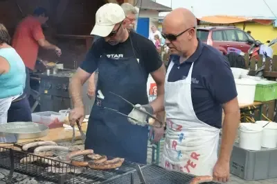 Zwei Männer grillen Würste auf einem Grill im Freien. Einer trägt einen Hut und eine Schürze, der andere eine Sonnenbrille. Ein Auto und Tische mit Eimern sind im Hintergrund.