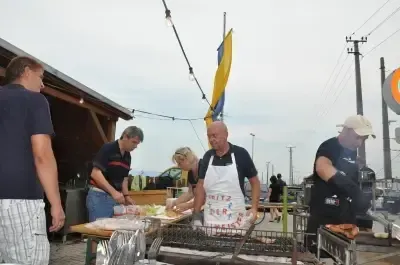 Eine Gruppe von Menschen bereitet Essen im Freien zu. Ein Mann in einer weißen Schürze grillt, während andere stehen und möglicherweise helfen. Es gibt Tische mit Essen und eine Fahne im Hintergrund.