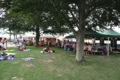Eine große Gruppe von Menschen versammelt sich in einem Park unter Zelten. Einige sitzen auf Bänken, während andere auf Decken liegen. Es gibt Bäume und offene Grasflächen um sie herum.