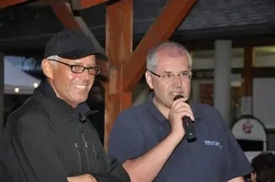 Zwei Männer stehen im Freien. Ein Mann lächelt, während der andere ein Mikrofon hält. Beide tragen Brillen und scheinen zu sprechen. Im Hintergrund befindet sich eine Holzkonstruktion.