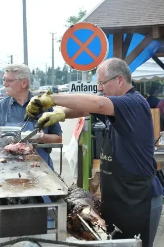 Zwei Männer kochen ein großes gebratenes Schwein auf einem Grill im Freien. Ein Mann schneidet Fleisch, während der andere beobachtet. Sie befinden sich in einem öffentlichen Bereich mit einem Schild und einem Gebäude im Hintergrund.