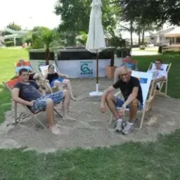 Eine Gruppe von Menschen sitzt auf Liegestühlen auf einem sandigen Bereich mit einem grünen Rasen, Bäumen und einem weißen Schirm im Hintergrund.