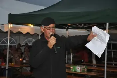 Ein Mann in einem schwarzen Jackett und mit einer Brille steht unter einem grünen Zelt, hält ein Mikrofon und liest von einem Papier. Im Hintergrund sitzen Menschen an Tischen.